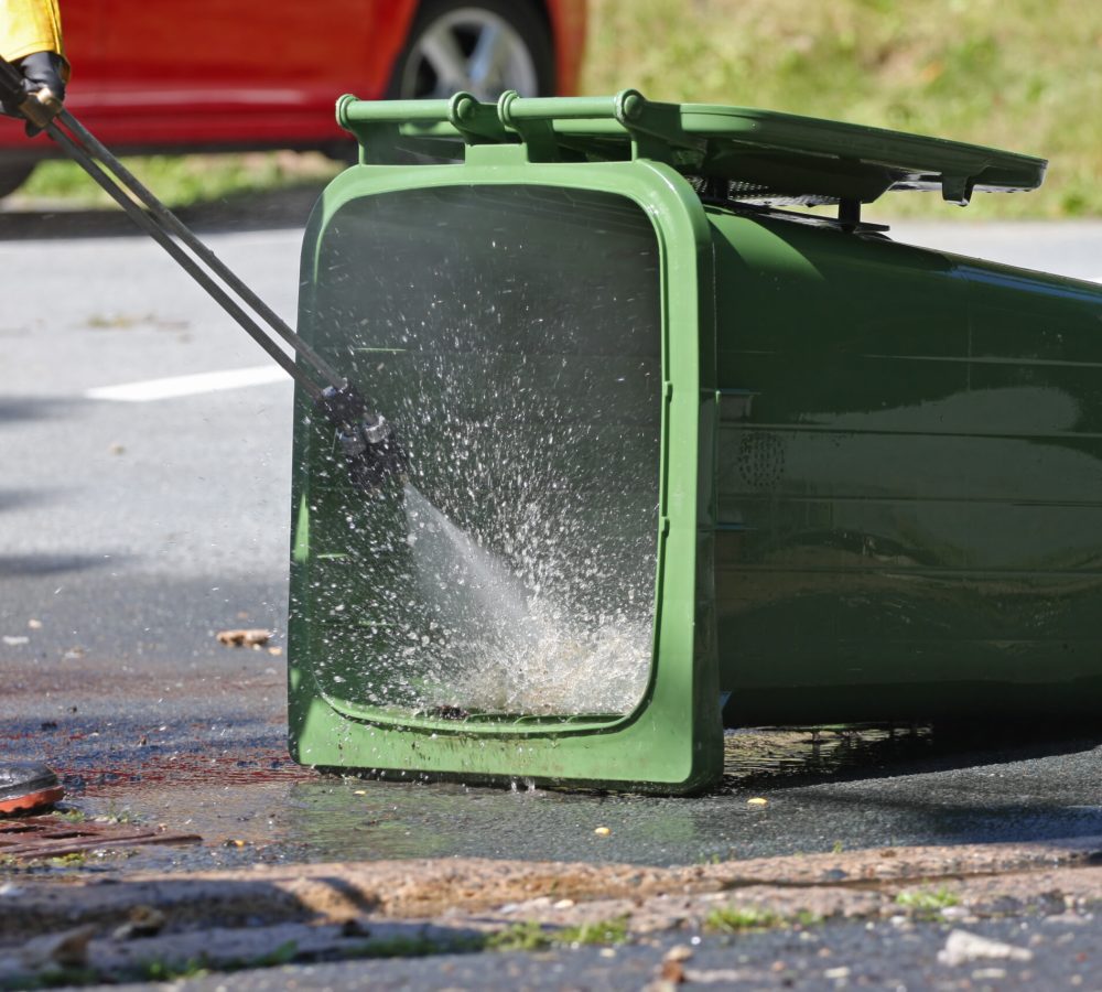 Sanitation worker uses a power washer to clean a compost recycling green bin.